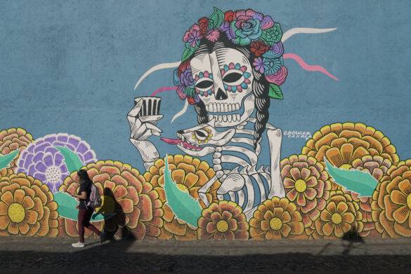 Graffiti von einem mexikanischen Totenkopf vor dem eine junge Frau mit Backpack durchläuft.